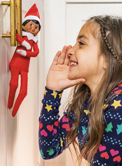 Scout Elf hanging from a cabinet handle. A young girl is leaned into the elf with her hand around her mouth telling the elf a secret.