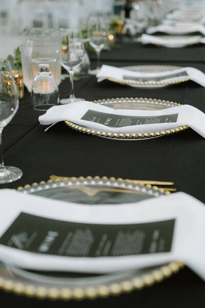 A sophisticated wedding table setting with black tablecloths, gold-rimmed glass chargers, white napkins, and modern black-and-white menus, complemented by elegant glassware and candlelight.