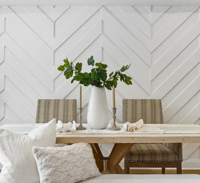 Elegant dining area with a white chevron accent wall, wooden table, neutral-toned chairs, and a white vase centerpiece with green foliage.