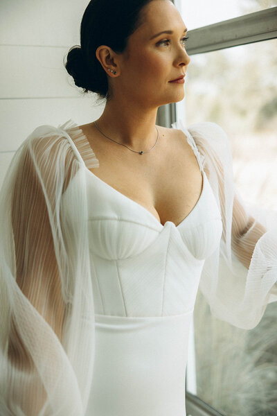 Bride in a modern wedding gown with sheer bishop sleeves gazing out the window, captured during a calm wedding morning in Austin, TX.