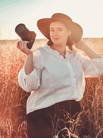 very warm sunset photo of Massachusetts Wedding Photographer Avid Aperture Photography wearing a long-sleeve white blouse and black wide-rim hat holding her camera in a tall grassy field located at Sapowet Marsh in Tiverton, Rhode Island