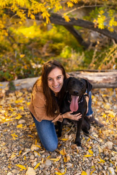 A woman with her black lab out for a hike in the woods during fall. The leaves are bright yellow.