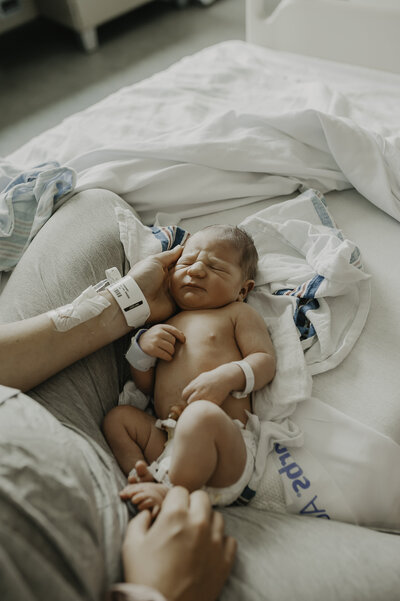 Verona B Photography captures a fresh newborn less than 48 hours old laying on the hospital bed between her mom's legs. The newborn is peacefully asleep and chunky and beautiful. 