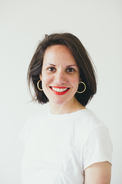 Kaya Ramjee with shoulder-length dark hair, wearing a white t-shirt and red lipstick, smiling in front of a white background.