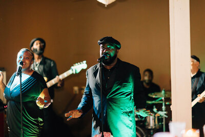 A band performs outdoors; a man in a dark jacket and hat sings into a microphone at center and musicians play guitar and drums in the background under warm lighting.