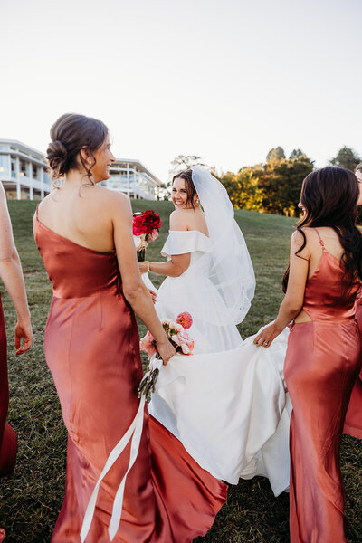 Bride looking her shoulder smiling at the camera while her bridesmaid help carry her dress. The Marion wedding venue shown in the background. Photo by Acacia and Grace Photography