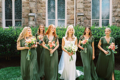 A bride wearing all white and a veil is surrounded by her five bridesmaids wearing green as they walk through a church courtyard in Northern Virginia