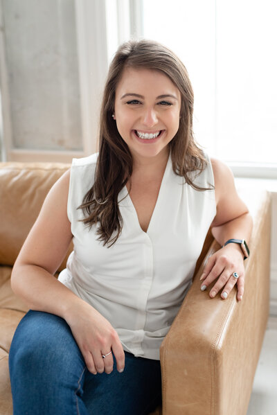 Bee smiles at the camera while sitting on a brown couch crossing her legs, she wears a casual outfit of jeans and a white button up blouse