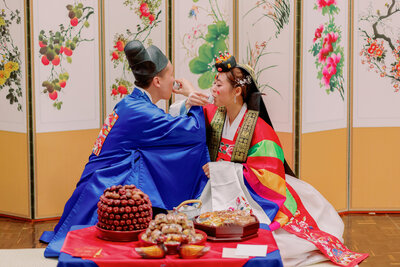 A newlywed couple sitting on the floor eating with their arms linked 