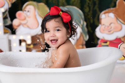 Child wearing a red bow bandana is seen splashing water in a white mini tub with various Snow White characters surrounding her.