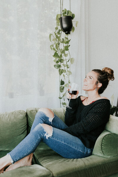 Woman in side profile as she looks up and has a glass of wine while sitting on a green sofa