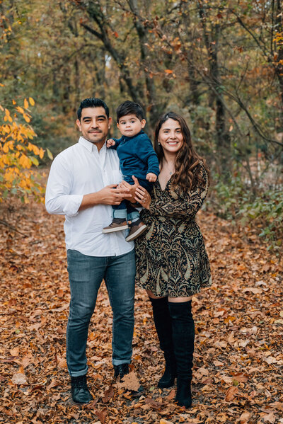Mom and dad hold baby in woods surrounded by fall leaves.