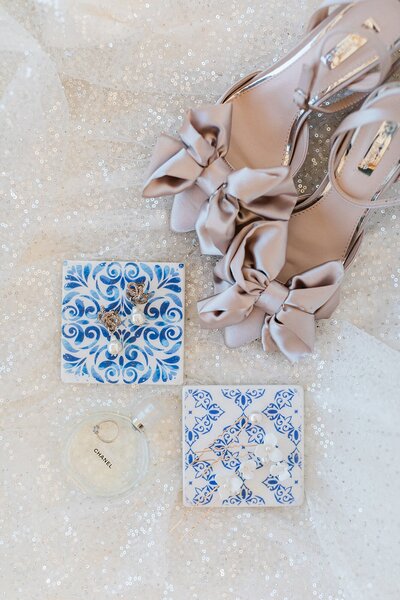 Flatlay of elegant bridal accessories including blush bow heels, wedding earrings, and Chanel perfume styled on decorative blue tiles and sparkly fabric