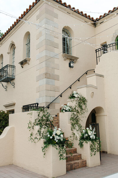 Exterior photo of a cream-colored Spanish Colonial or Mediterranean style building featuring a stone staircase decorated with climbing ivy and white white floral arrangements. 