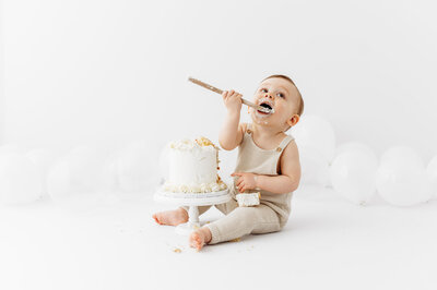 Baby boy with a white cake, wearing beige romper and holding a wooden spoon