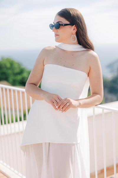 A bride stands in the foreground in a white gown, looking to the side, while a man ascends a staircase in the background, within an arched doorway. This scene captured by a luxury wedding