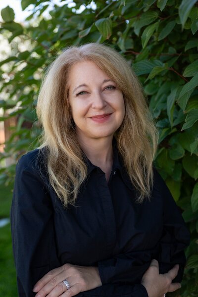 Cyndi Stewart smiling woman with long, wavy hair sits comfortably on a couch, wearing a teal top and jewelry, in a welcoming and relaxed setting.