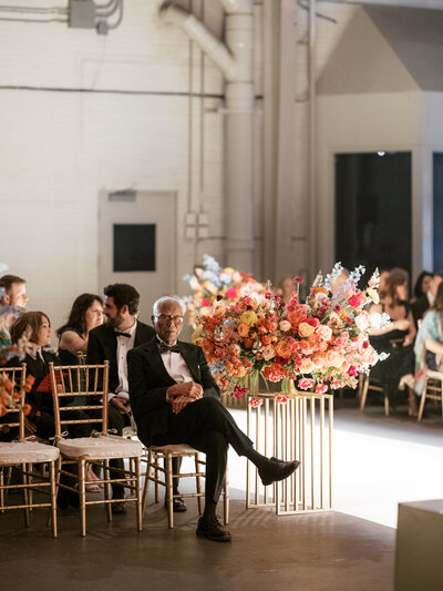 April M Payne Photography captured a wedding ceremony scene with a guest sitting alone, surrounded by a vibrant floral arrangement and gold chairs, while others are in the background, creating a warm and intimate atmosphere.