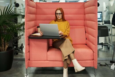 A professional woman sitting in a modern pink cushioned booth, working on a laptop, conveying focus and a stylish work environment.