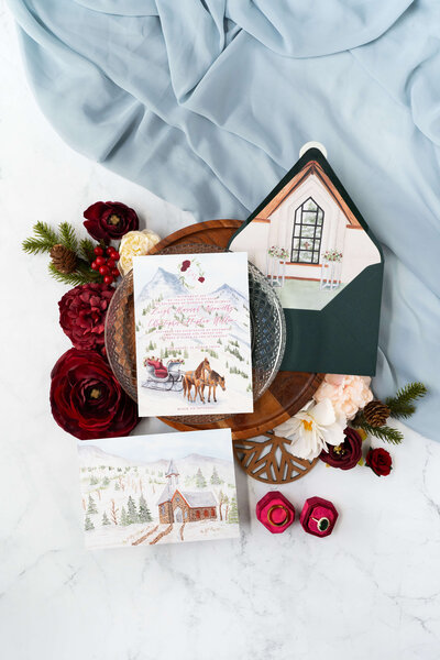 Invitation flatlay for luxury Beaver Creek winter wedding, highlighting watercolor horse-drawn carriage and full-art chapel painting on formal invitation, with dark green envelope and  an envelope liner of the chapel interior.