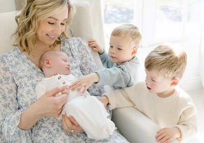 mom sitting and holding newborn baby while older twin brothers look on.