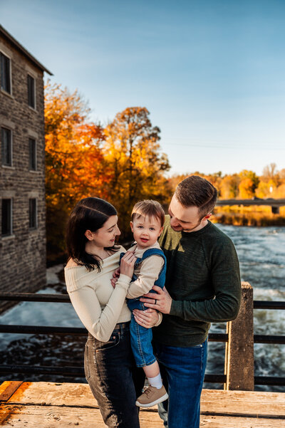 Young family smiling with their toddler at Watson’s Mill in Manotick during Ottawa fall mini session, colorful autumn trees in the background.