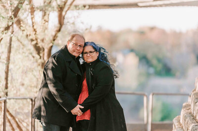Husband stands behind wife with his arms around her.