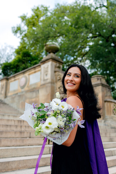 Graduation portrait of a student holding flowers and gown while smiling at Bethesda Terrace in NYC.
