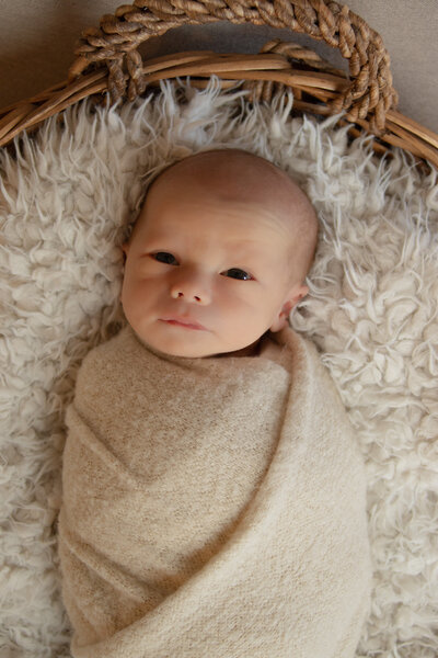 newborn boy in tan swaddle looking up at the camera in basket on floor