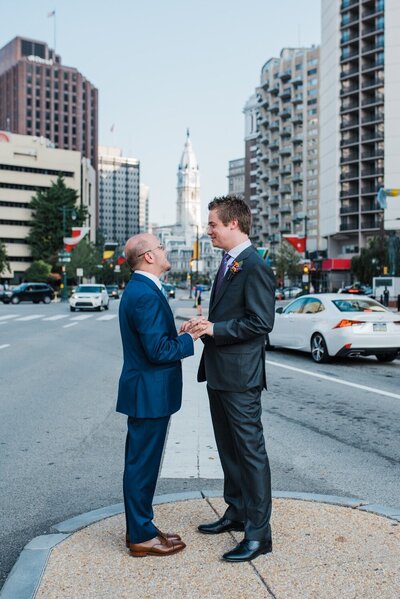 Two grooms in blue and gray suits standing holding hands staring into each other’s eyes with the city skyline outback.