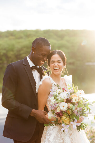 Groom and bride laugh in front of New York lake