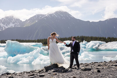 Bride and groom pose on the rocky shore of an Alaskan glacial lake, surrounded by floating icebergs and towering mountains. The bride’s white wedding dress flows as she steps onto a rock, holding her groom’s hand, capturing the adventurous and scenic beauty of an Alaska elopement.