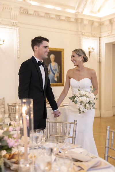 A couple in wedding attire holds hands and smiles at each other in an elegant, ornate room. The bride holds a bouquet of white flowers, and the groom wears a black tuxedo. Tables are set with glassware and flowers in the background.