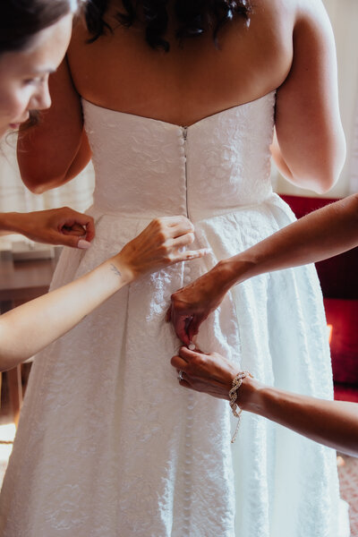 Two bridesmaids stand behind bride wearing elegant strapless gown as they help button the buttons lining the back.