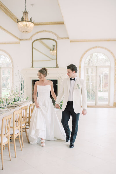 Bride and groom walking through the wedding breakfast room looking at the table decorations. They are holding hands and bride is wearing a white strapless ballgown and the groom has a white tuc jacket and black trousers and bowtie.