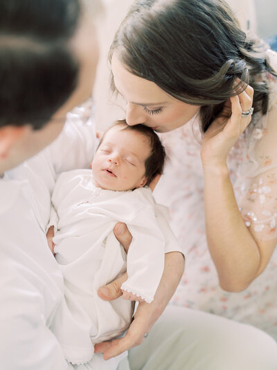 A brown-haired mother leans down to kiss her brown-haired baby's head while her husband holds her during their Arlington, Virginia newborn session.