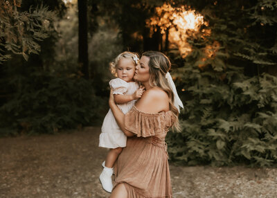 A mother in a pretty pink dress squeezing and kissing her toddler girl.