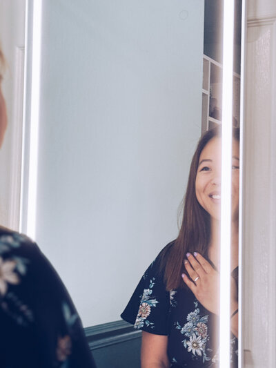 An Asian-American woman with her hand on her chest and smiling at her reflection.