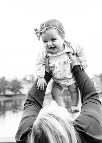 Black and white photo of baby girl held in the air by mom, smiling and giggling