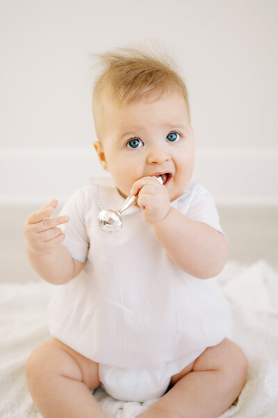 Baby boy in white romper chews on silver rattle during baby photo session in Raleigh