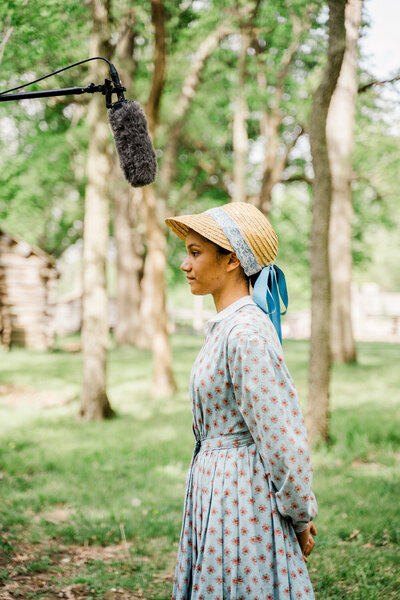 an interview subject dressed in period  attire stands under a boom mic on a storyteller studios set in a historic location
