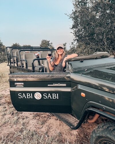 A woman in a safari jeep, glass of wine in hand, soaking in the wild beauty around her with effortless elegance.