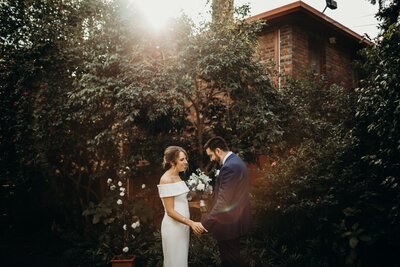 A bride and groom in front of a tree, holding hands sharing an intimate moment after a first look with the afternoon sun behind them