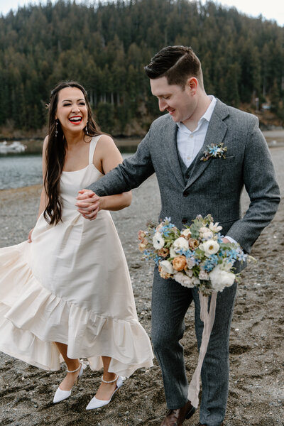 Elopement couple holds hands smiling on Seldovia beach with bouquet.