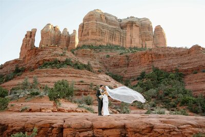 Couple standing at the base of Cathedral Rock in Sedona AZ taking their bridal portrait and the brides' veil is blowing in the wind