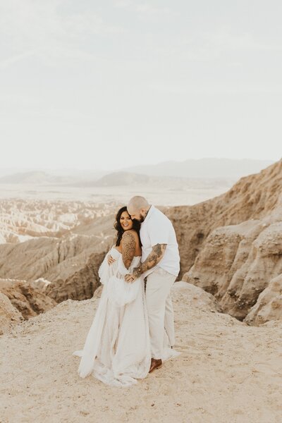 husband hugs wife from behind for. their maternity photos in the mountains