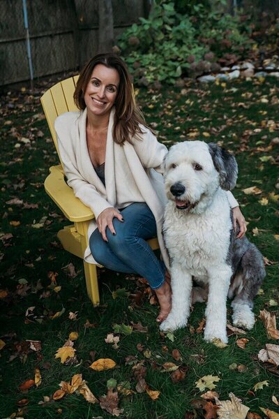 Gina Cooperman sitting outdoors with a large dog, surrounded by autumn leaves.
