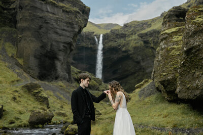 Romantic elopement photo at Kvernufoss waterfall in Iceland photographed by Destination Elopement Photographer — groom in a black suit kisses the bride’s hand as she smiles in a flowing tulle dress, surrounded by moss-covered cliffs and cascading falls.