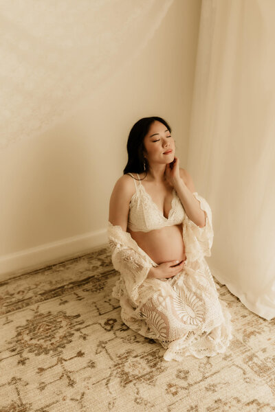 Expecting mother wearing a cream colored bralette and kimono while kneeling on the ground for a studio portrait in OKC.