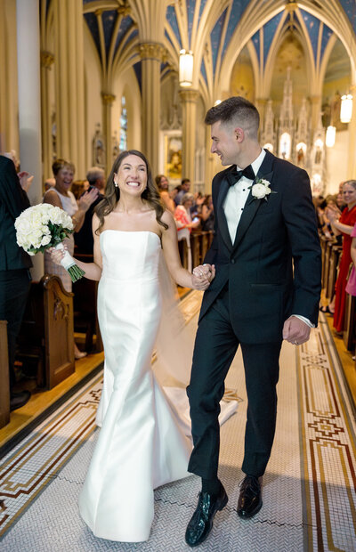 A bride in a white gown and a groom in a black tuxedo walk hand-in-hand down the aisle of a church, smiling joyfully. The churchs gothic architecture and stained glass can be seen in the background, with guests seated and watching.